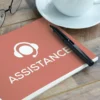 Un cahier de couleur saumon portant le logo "Assistance" est posé sur ce bureau en bois, avec des lunettes, un stylo et une tasse, reflétant les services d'assistant virtuel offerts à Trois-Rivières.