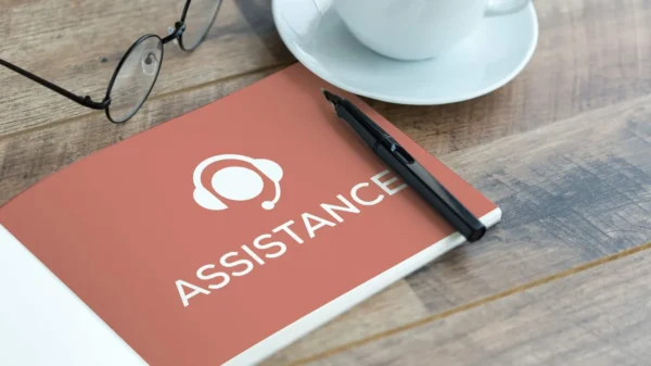 Un cahier de couleur saumon portant le logo "Assistance" est posé sur ce bureau en bois, avec des lunettes, un stylo et une tasse, reflétant les services d'assistant virtuel offerts à Trois-Rivières.